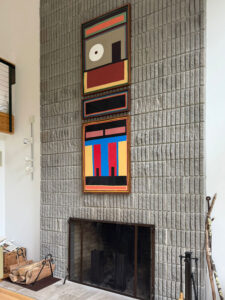 Triptych art installation on a textured gray concrete block fireplace wall, featuring bold geometric paintings in vibrant colors.